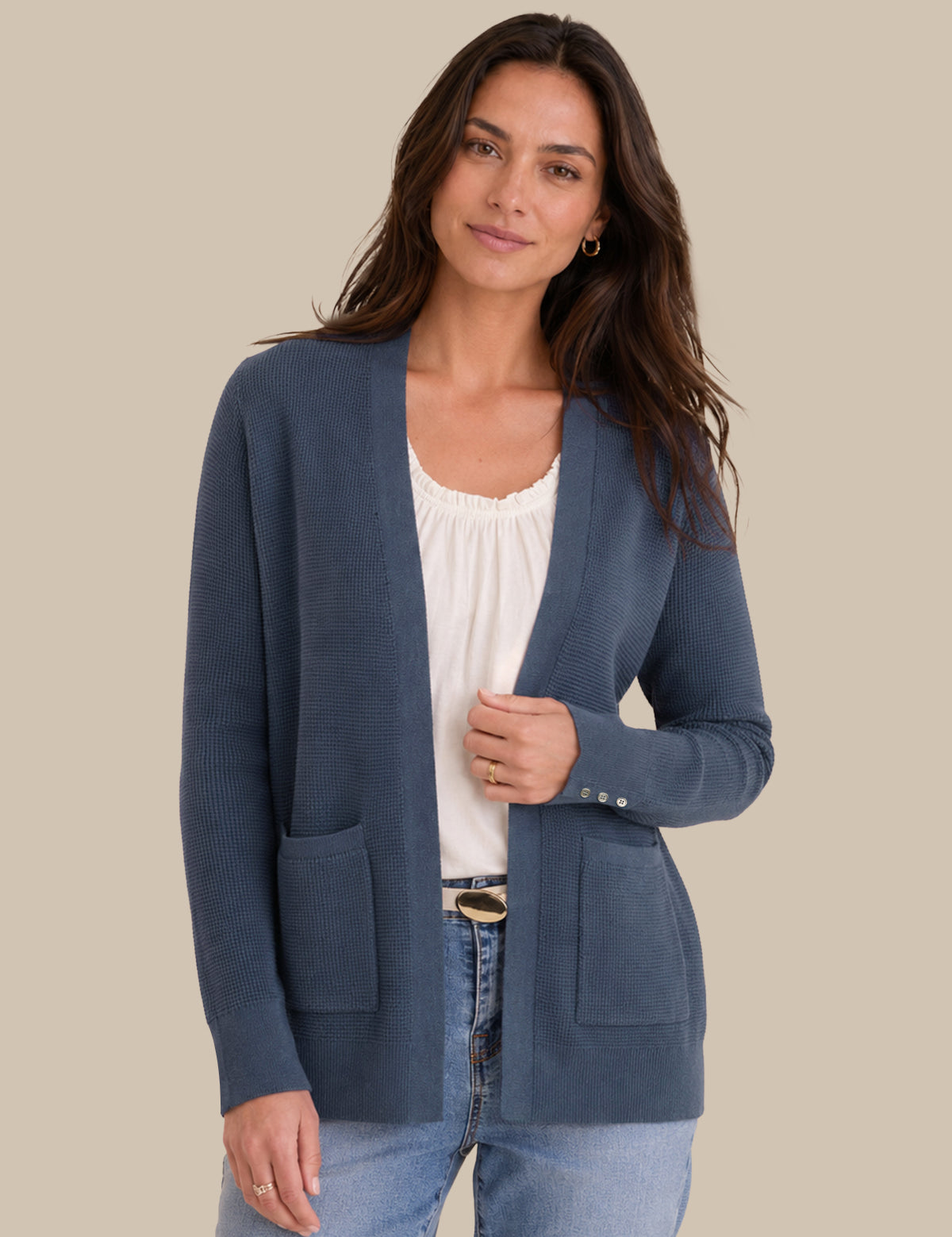 A woman modeling a slate blue open-front cardigan with front patch pockets over a white top and light-wash jeans, set against a solid beige background.