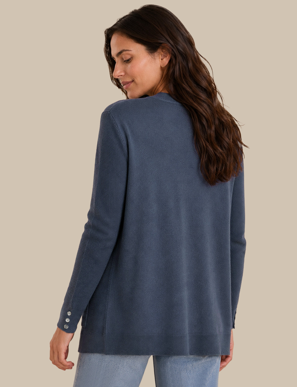 A woman with long dark brown hair is shown from the back wearing a slate blue long-sleeve knit top with three decorative buttons on the cuff, paired with light blue jeans, against a solid beige background.