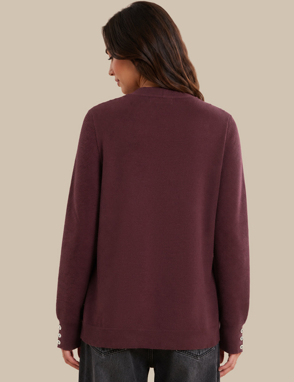 A woman shown from the back wearing a long-sleeve knit sweater in a deep plum or burgundy shade, paired with dark jeans, against a solid beige background.