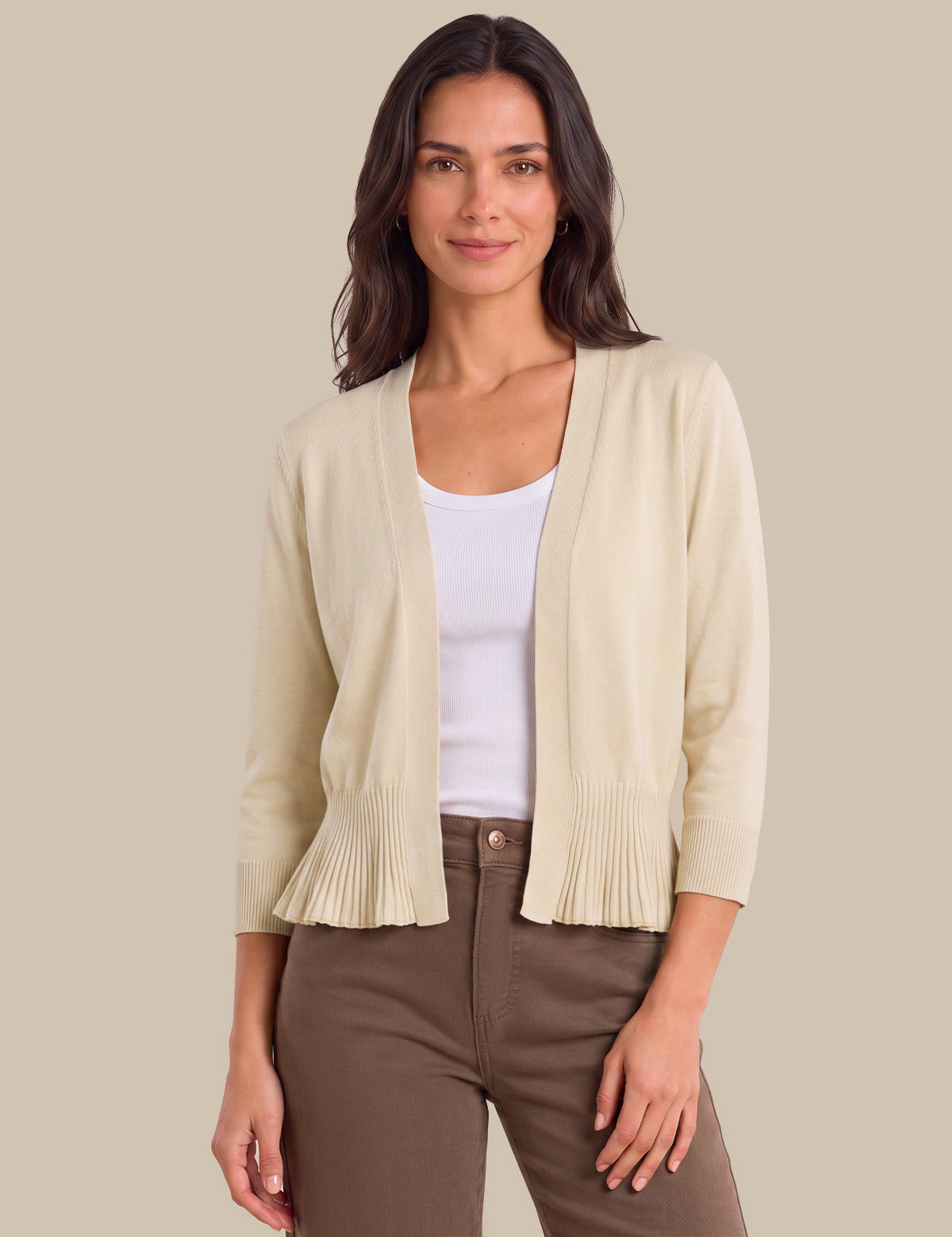 A woman modeling a light beige open-front cardigan with pleated ruffle trim at the hem and cuffs, worn over a white tank top and brown pants, against a solid beige background.