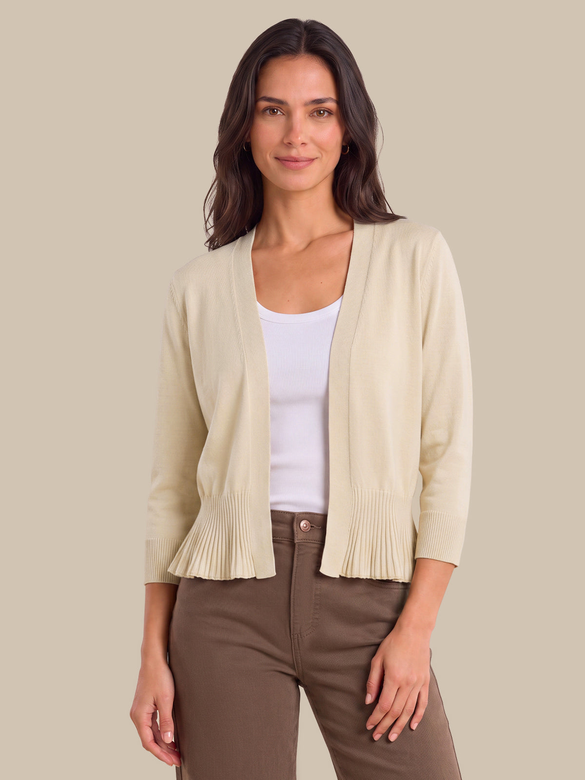 A woman modeling a light beige open-front cardigan with pleated ruffle trim at the hem and cuffs, worn over a white tank top and brown pants, against a solid beige background.