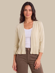 A woman modeling a light beige open-front cardigan with pleated ruffle trim at the hem and cuffs, worn over a white tank top and brown pants, against a solid beige background.