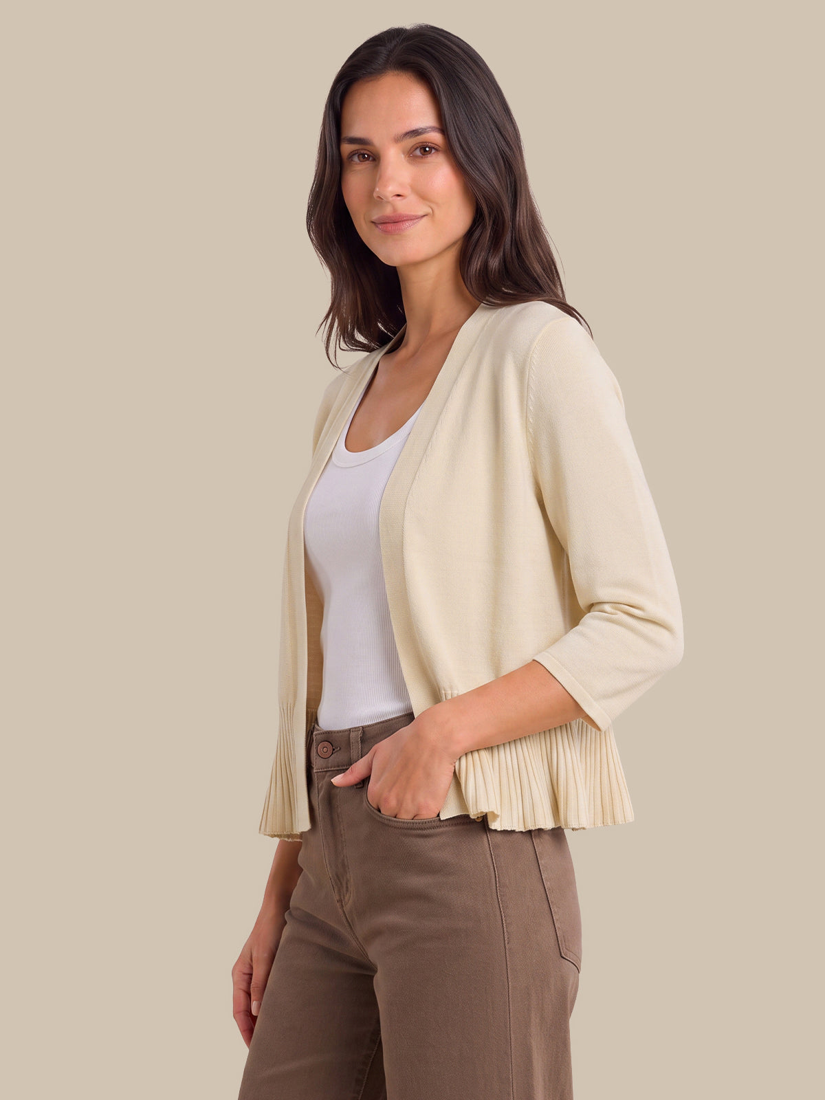 A woman modeling a cream-colored open-front cardigan with pleated fringe at the hem, worn over a white tank top and brown pants, against a solid beige background.