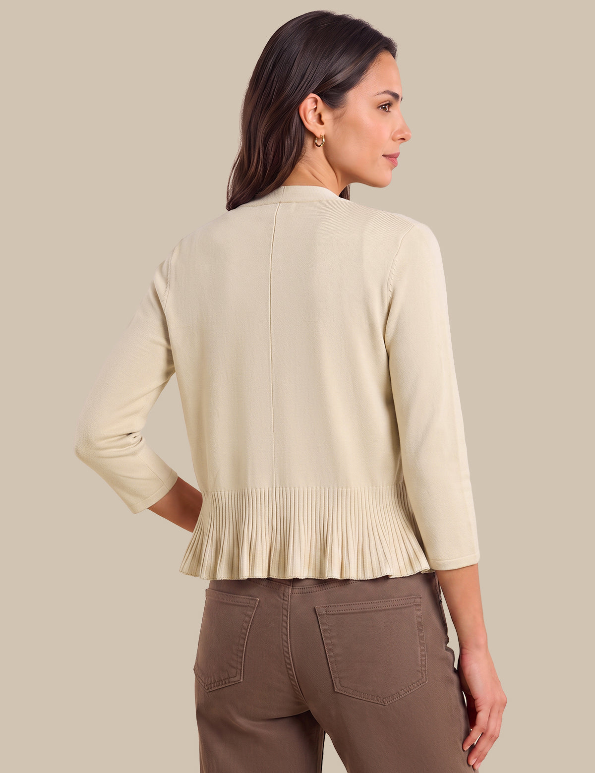 A woman shown from the back wearing a light beige cropped cardigan with three-quarter sleeves and a pleated hem, paired with brown pants, against a solid beige background.