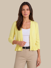 A woman modeling a pale yellow open-front cardigan with pleated cuffs and hem, worn over a white tank top and beige pants against a solid beige background.