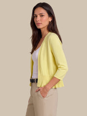 A woman modeling a light yellow knit cardigan with three-quarter sleeves and a pleated hem, worn over a white top and beige trousers, against a solid beige background.