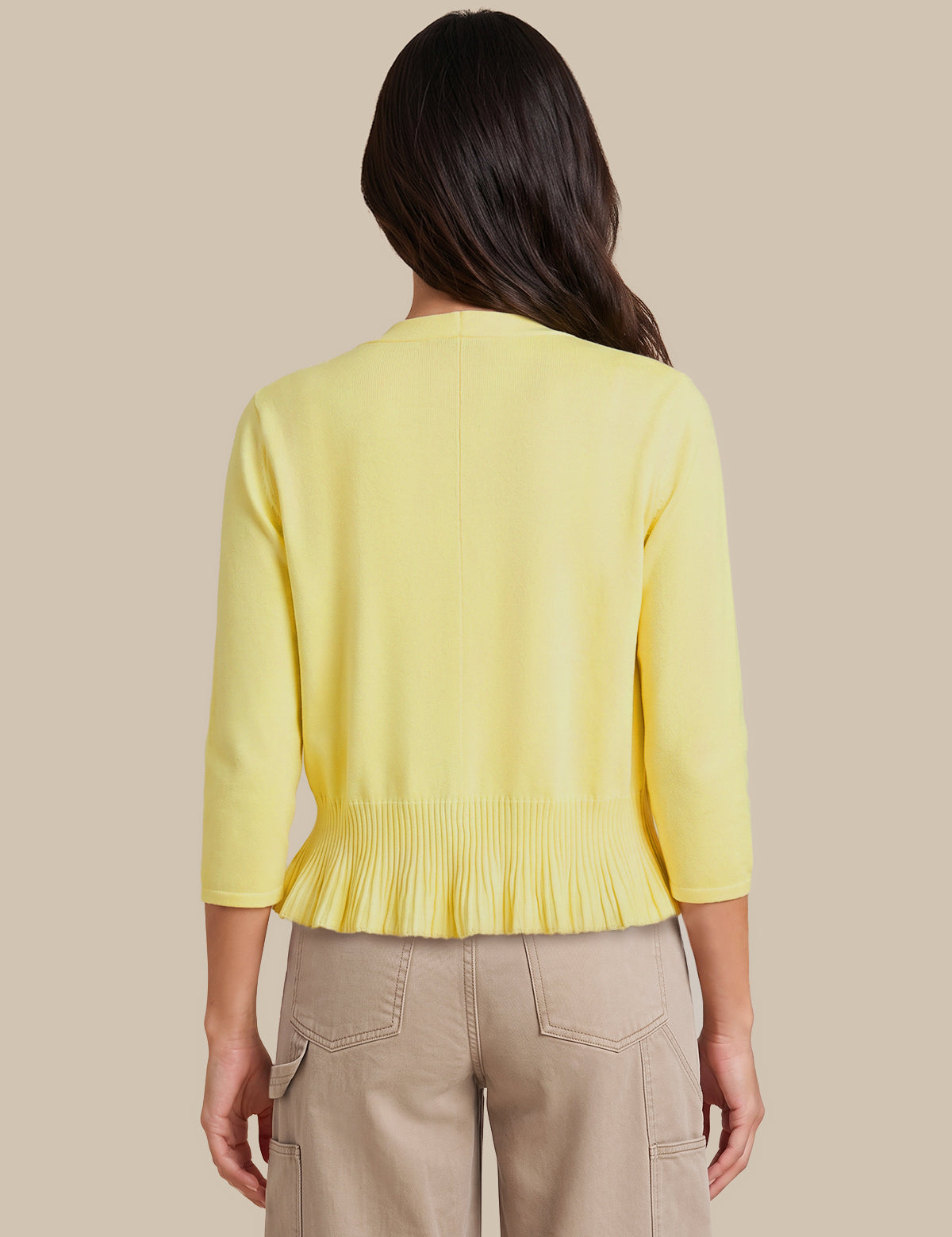 A woman viewed from the back wearing a pale yellow knit cardigan with three-quarter sleeves and a gathered pleated hem, paired with beige cargo pants, against a solid beige background.