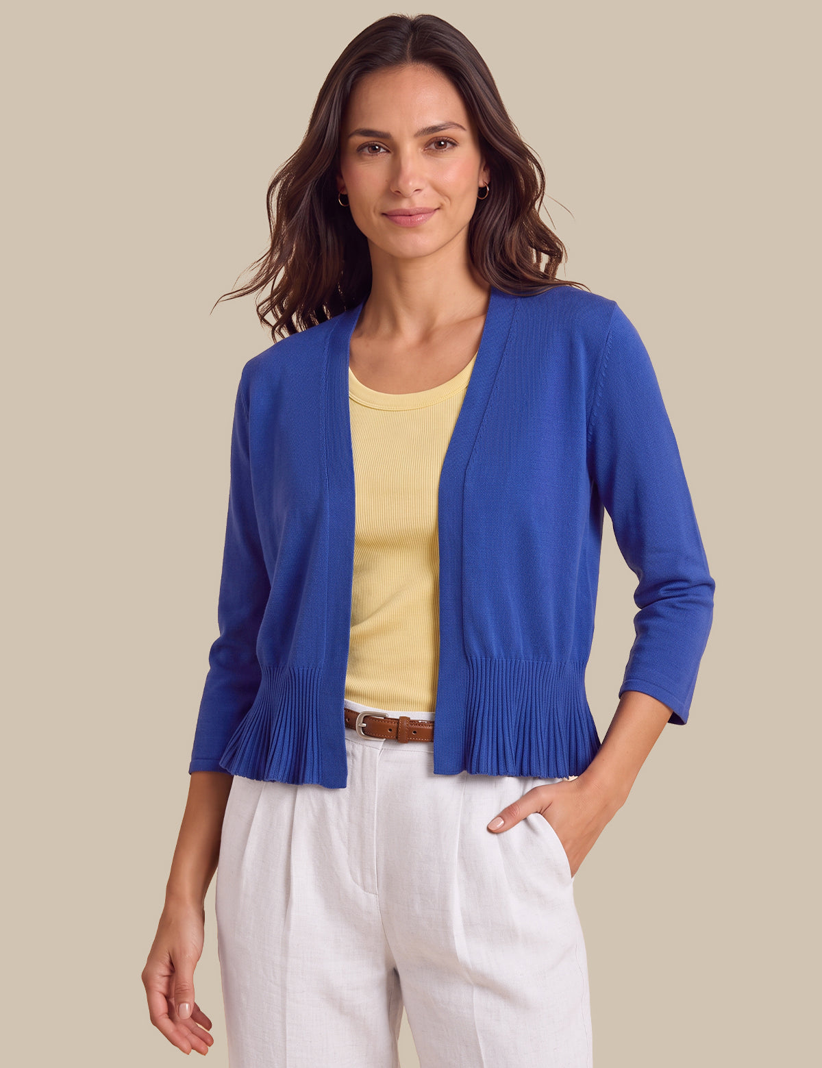 A woman modeling a royal blue open-front cardigan with pleated trim at the hem, worn over a pale yellow top and white pants against a solid beige background.