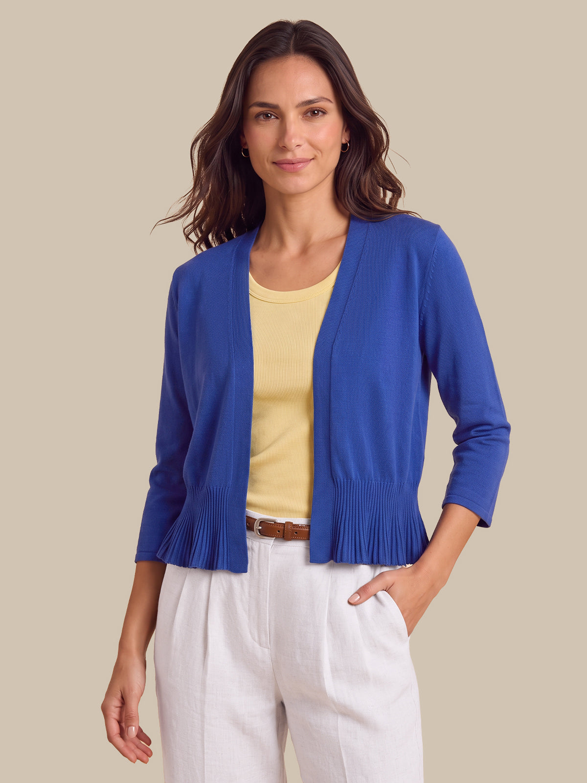 A woman modeling a royal blue open-front cardigan with pleated trim at the hem, worn over a pale yellow top and white pants against a solid beige background.