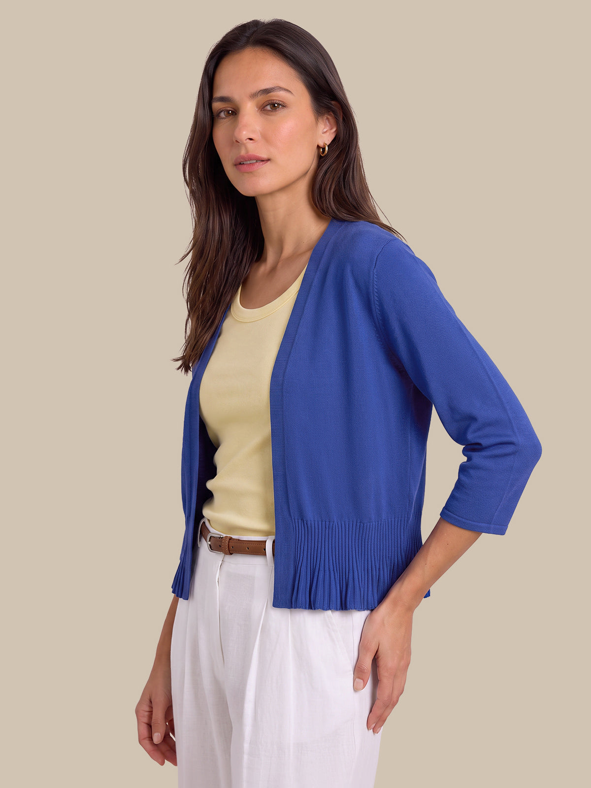 A woman modeling a royal blue open-front cardigan with three-quarter sleeves and a gathered, pleated hem, worn over a pale yellow top and white trousers against a solid beige background.