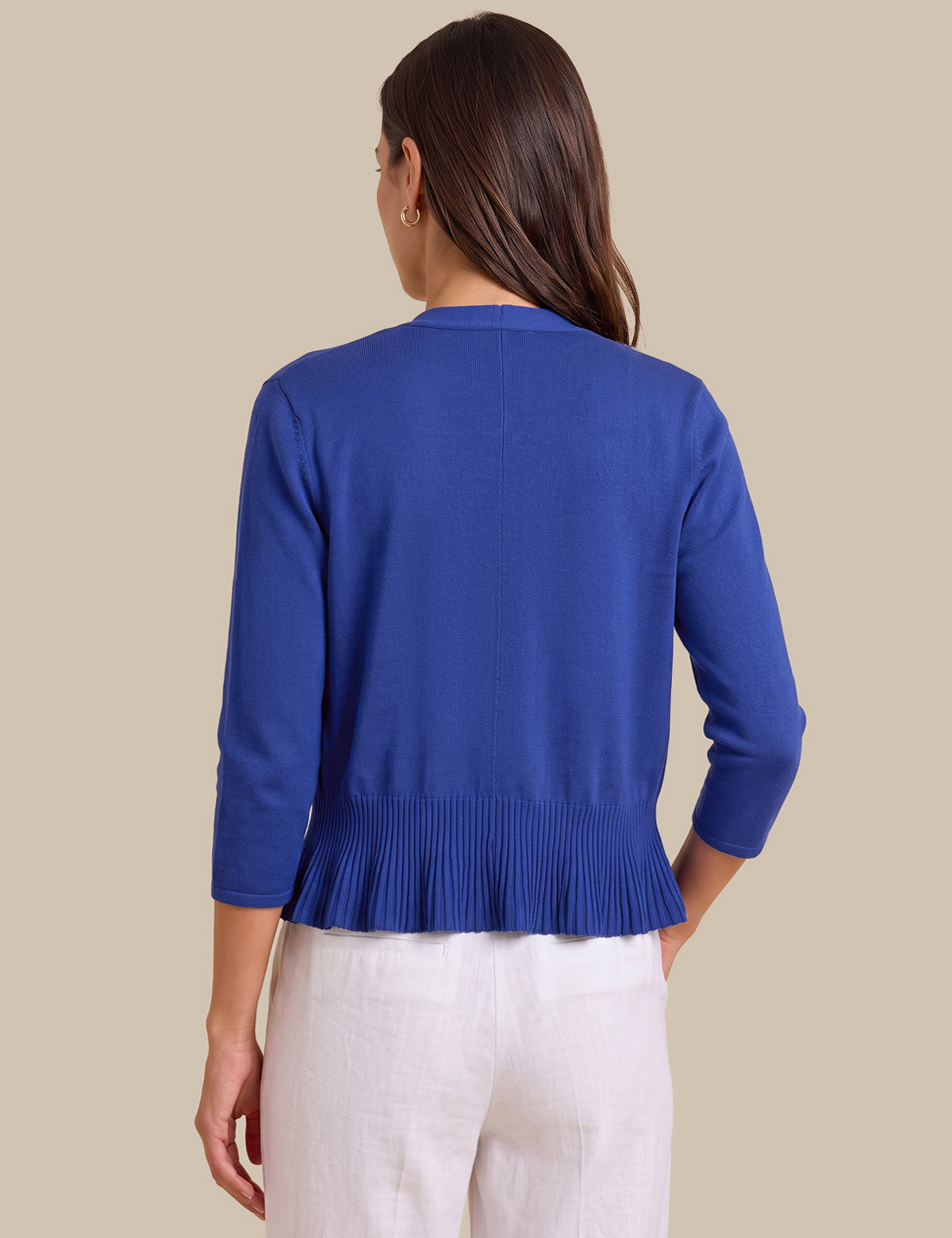 A woman shown from the back wearing a royal blue knit cardigan with three-quarter sleeves and a pleated hem, paired with white pants, against a solid beige background.