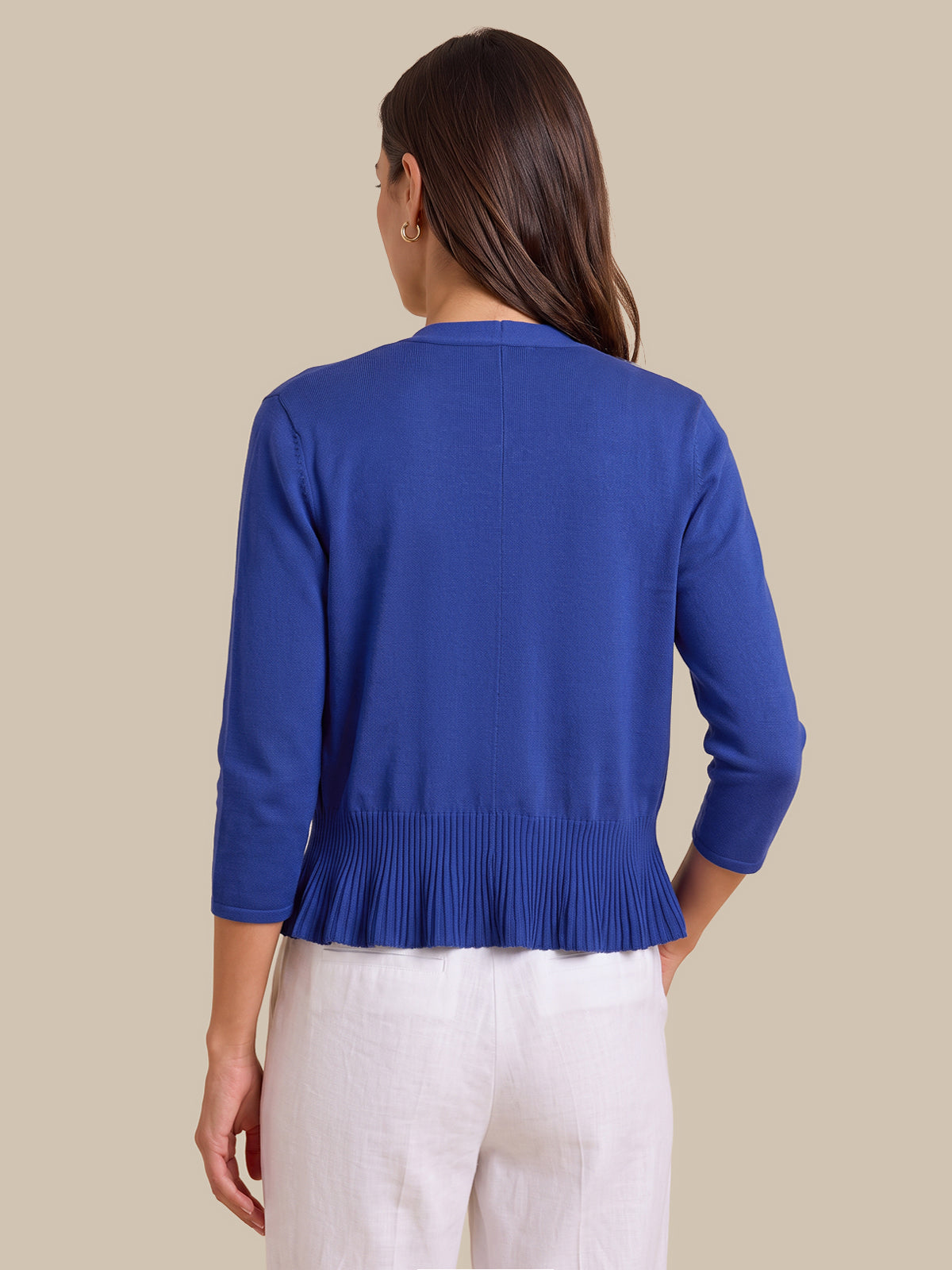 A woman shown from the back wearing a royal blue knit cardigan with three-quarter sleeves and a pleated hem, paired with white pants, against a solid beige background.