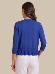 A woman shown from the back wearing a royal blue knit cardigan with three-quarter sleeves and a pleated hem, paired with white pants, against a solid beige background.