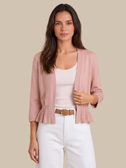 A woman modeling a dusty pink open-front cardigan with pleated ruffle details at the hem and cuffs, worn over a white tank top and white pants against a solid beige background.