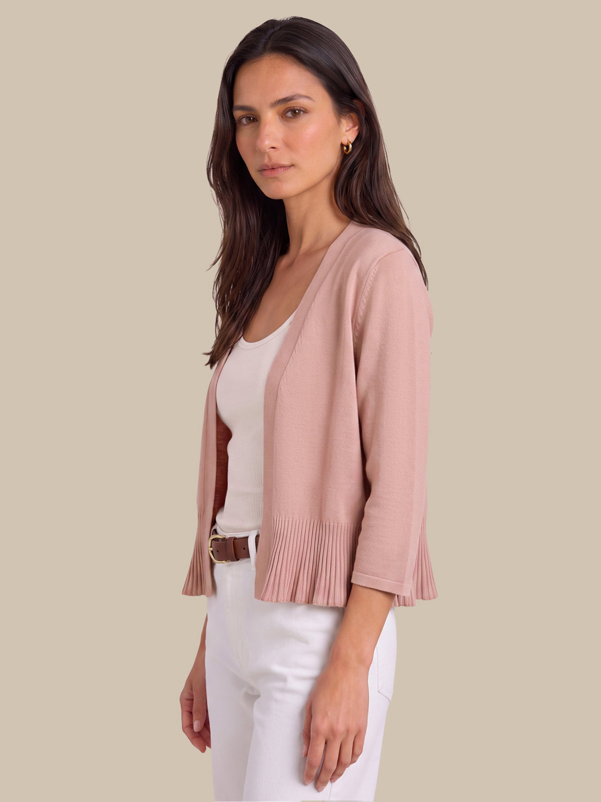 A woman modeling a light pink knit cardigan with pleated ruffle trim at the hem and three-quarter sleeves, worn over a white top and white pants against a solid beige background.