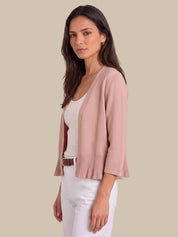 A woman modeling a light pink knit cardigan with pleated ruffle trim at the hem and three-quarter sleeves, worn over a white top and white pants against a solid beige background.
