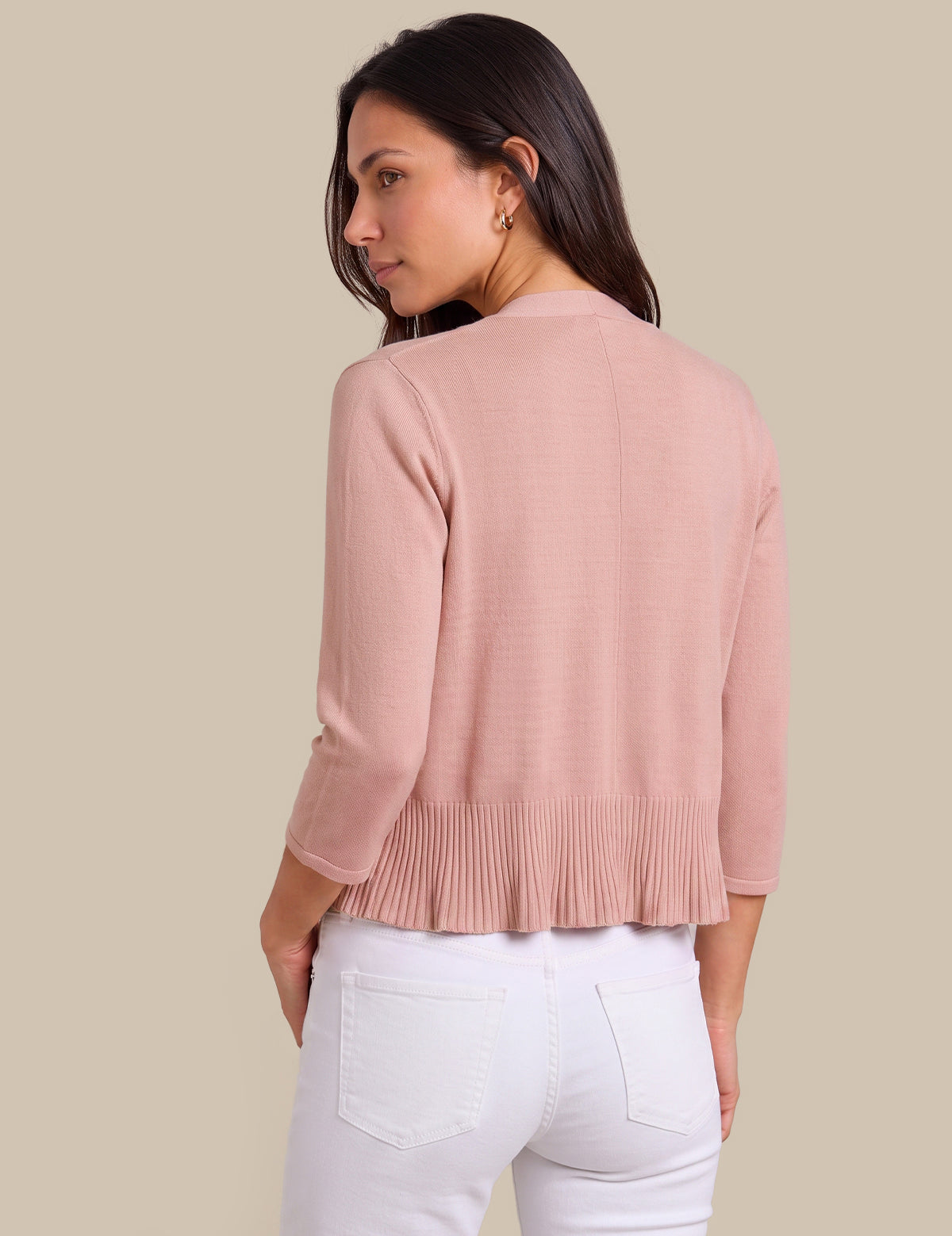 A woman shown from the back wearing a light pink three-quarter sleeve cropped cardigan with a pleated hem, paired with white pants, against a solid beige background.
