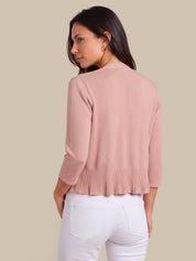 A woman shown from the back wearing a light pink three-quarter sleeve cropped cardigan with a pleated hem, paired with white pants, against a solid beige background.