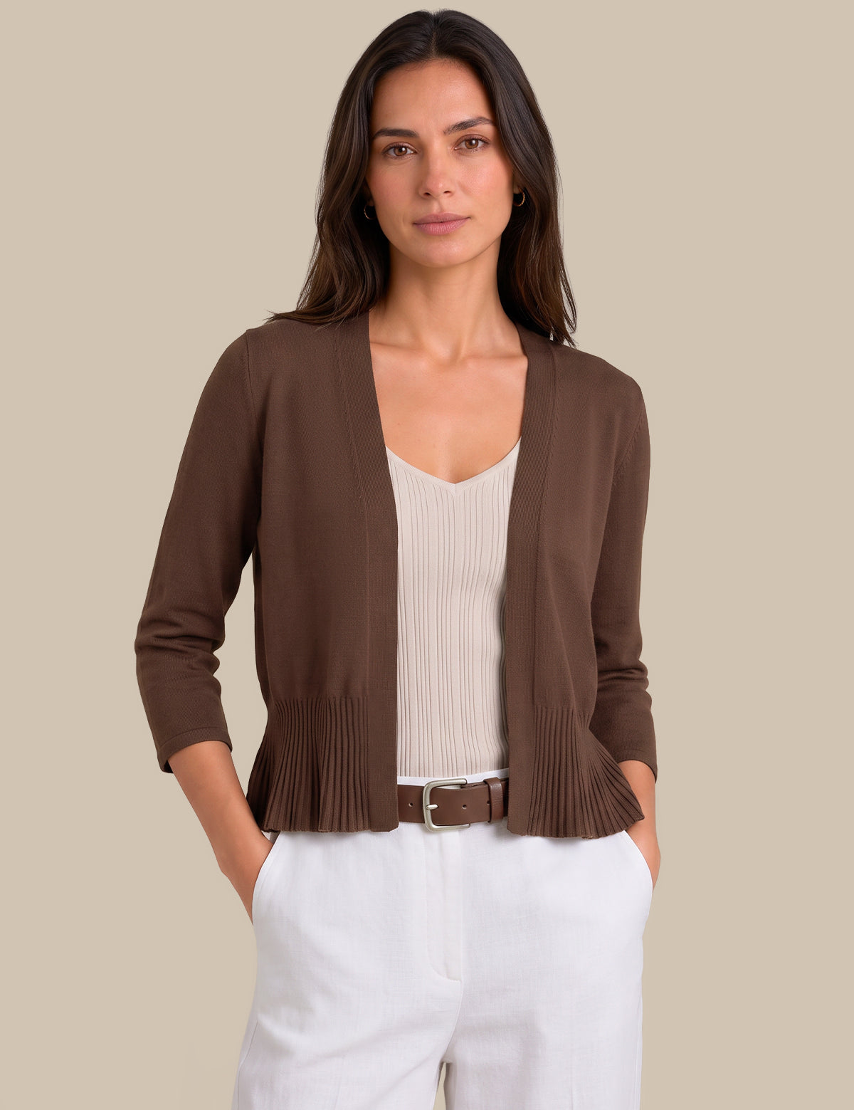 A woman modeling a brown open-front cardigan with pleated detailing at the hem, worn over a cream ribbed top and white pants against a solid beige background.