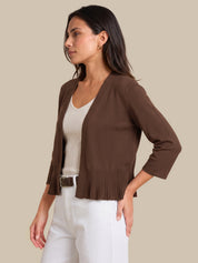 A woman wearing a brown open-front cardigan with pleated detailing at the hem and three-quarter sleeves, styled over a white ribbed top and white pants, against a solid beige background.