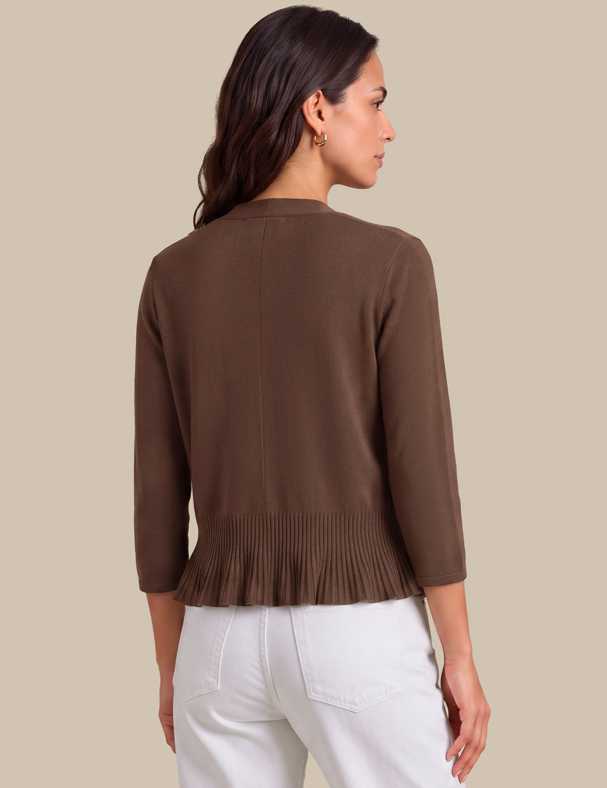 A woman shown from the back wearing a brown knit cardigan with three-quarter sleeves and a pleated hem, paired with white pants, against a solid beige background.