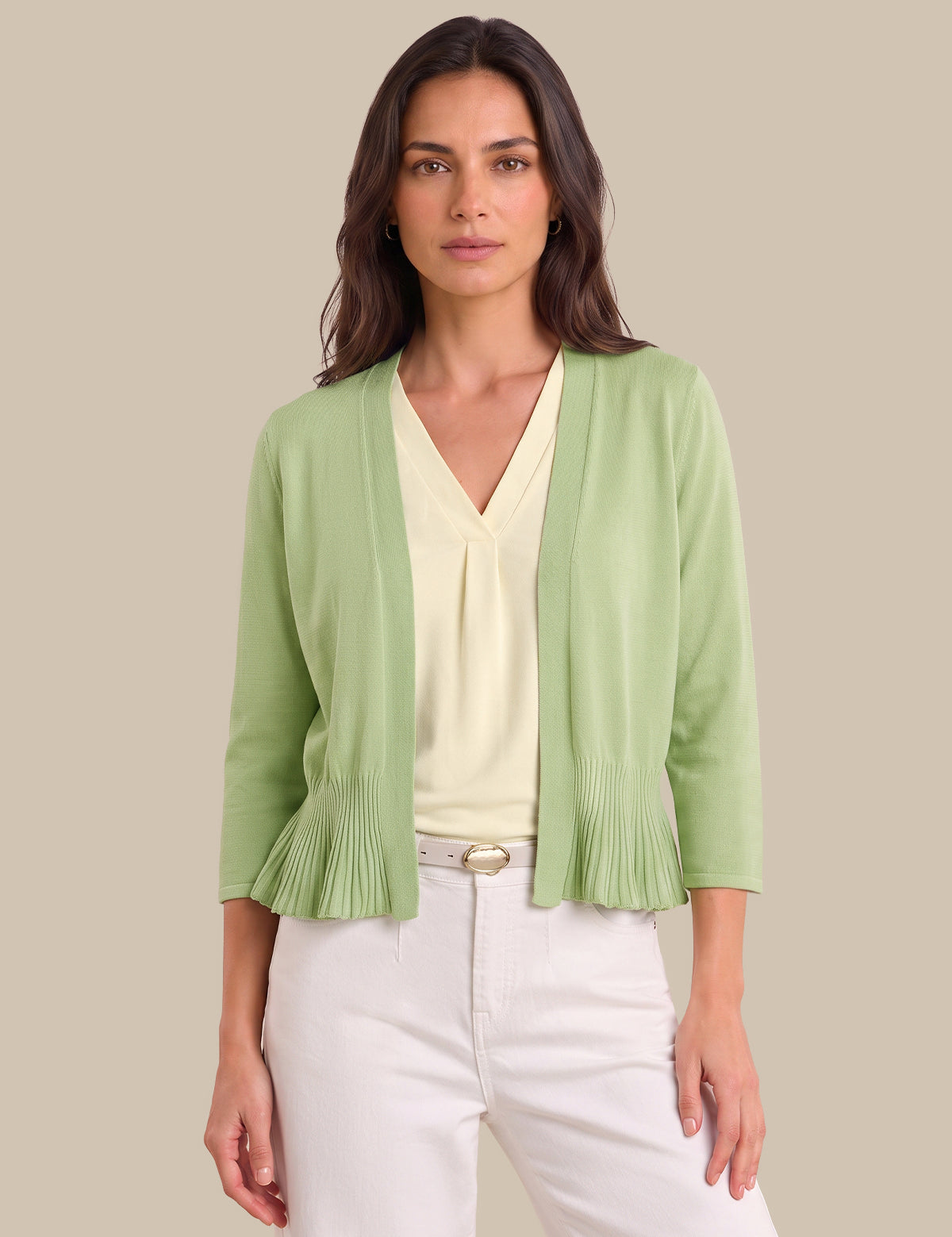 A woman modeling a light green open-front cardigan with pleated ruffle details at the hem and sleeves, worn over a cream V-neck top and white pants against a solid beige background.