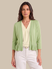 A woman modeling a light green open-front cardigan with pleated ruffle details at the hem and sleeves, worn over a cream V-neck top and white pants against a solid beige background.