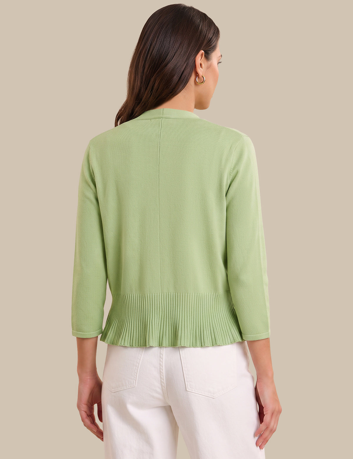 A woman shown from the back wearing a light sage green knit cardigan with three-quarter sleeves and a pleated hem, paired with white pants, against a solid beige background.