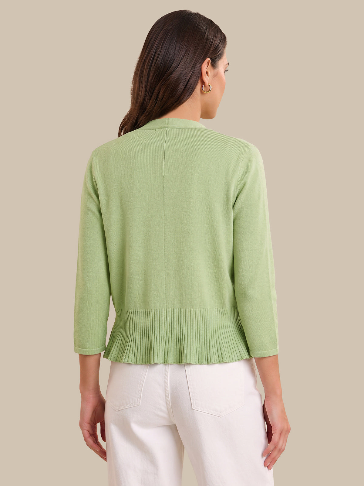 A woman shown from the back wearing a light sage green knit cardigan with three-quarter sleeves and a pleated hem, paired with white pants, against a solid beige background.