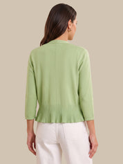 A woman shown from the back wearing a light sage green knit cardigan with three-quarter sleeves and a pleated hem, paired with white pants, against a solid beige background.
