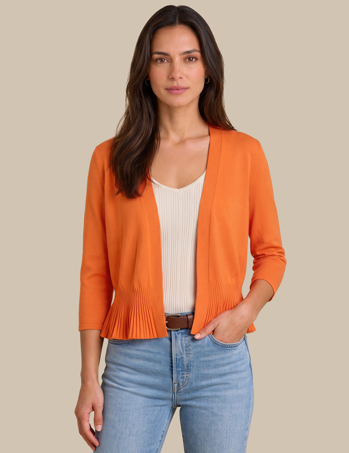 A woman modeling a bright orange open-front cardigan with three-quarter sleeves and a pleated hem, worn over a cream ribbed top and blue jeans against a solid beige background.
