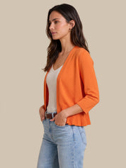 A woman wearing a bright orange knit cardigan over a white top and light blue jeans, standing against a solid beige background.