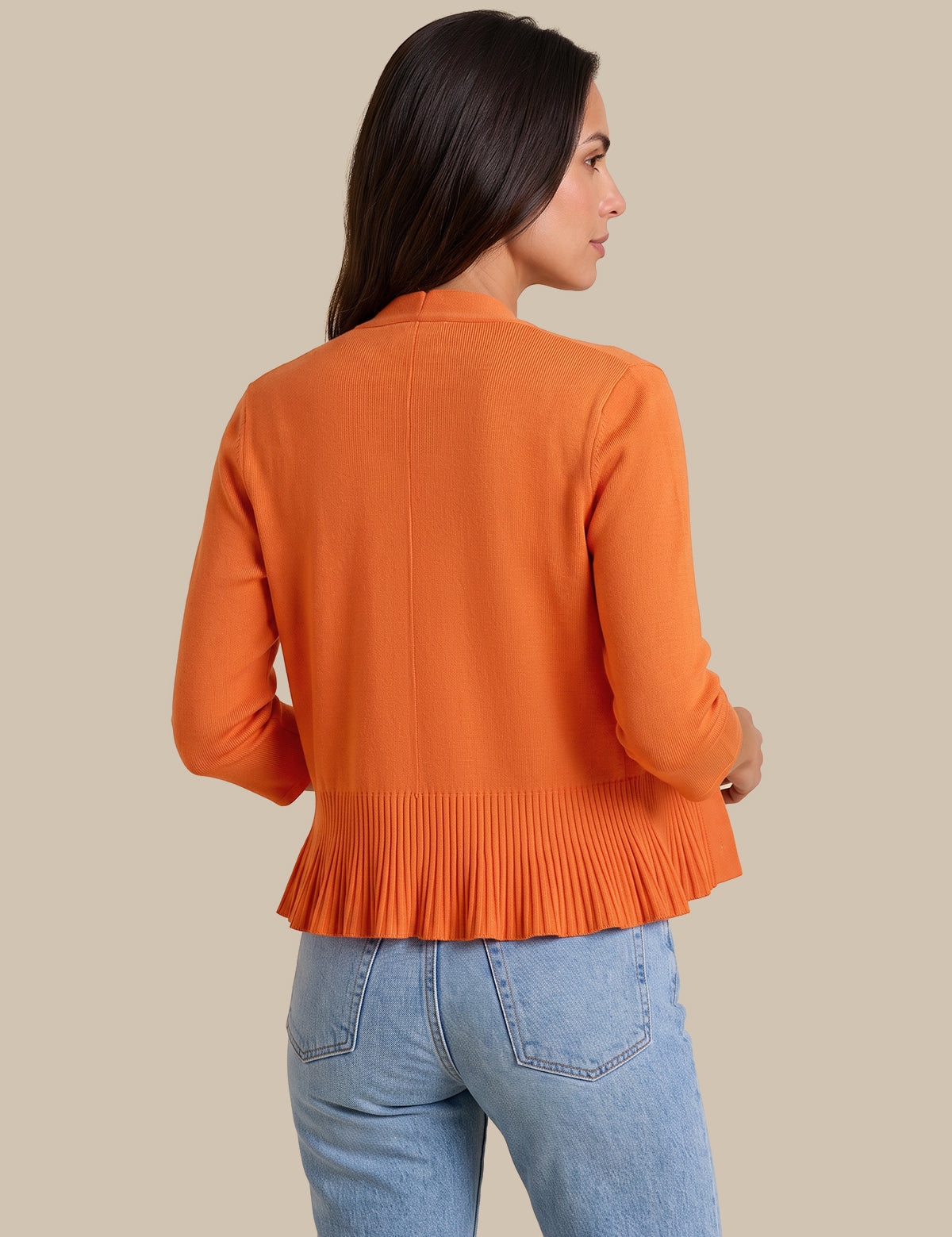 A woman shown from the back wearing a bright orange long-sleeve knit top with a pleated peplum hem, paired with light blue jeans, against a solid beige background.