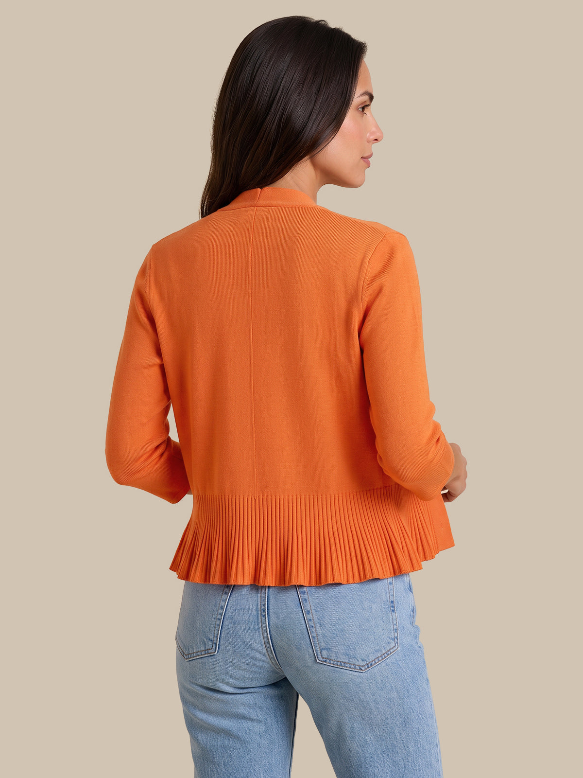 A woman shown from the back wearing a bright orange long-sleeve knit top with a pleated peplum hem, paired with light blue jeans, against a solid beige background.