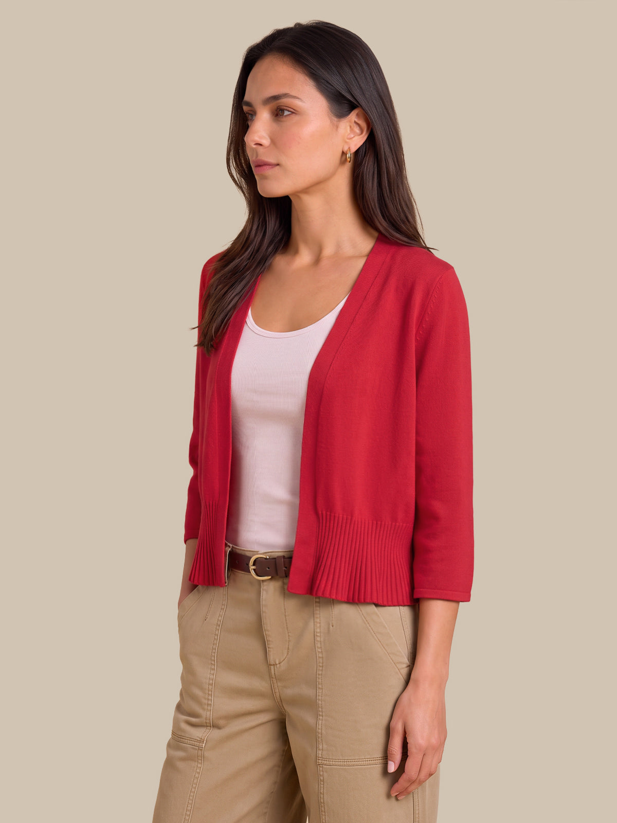 A woman modeling a bright red open-front knit cardigan with three-quarter sleeves, worn over a light pink tank top and beige pants, against a solid beige background.