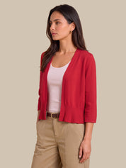 A woman modeling a bright red open-front knit cardigan with three-quarter sleeves, worn over a light pink tank top and beige pants, against a solid beige background.
