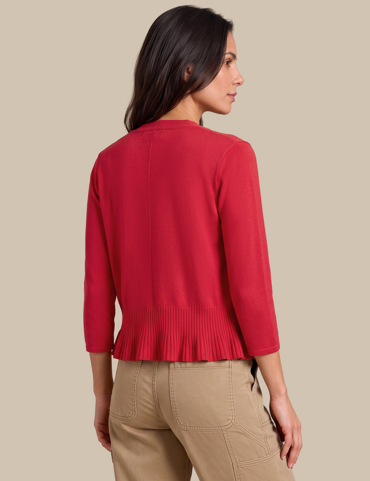 A woman shown from the back wearing a bright red knit cardigan with three-quarter sleeves and a pleated hem, paired with beige cargo pants, against a solid beige background.