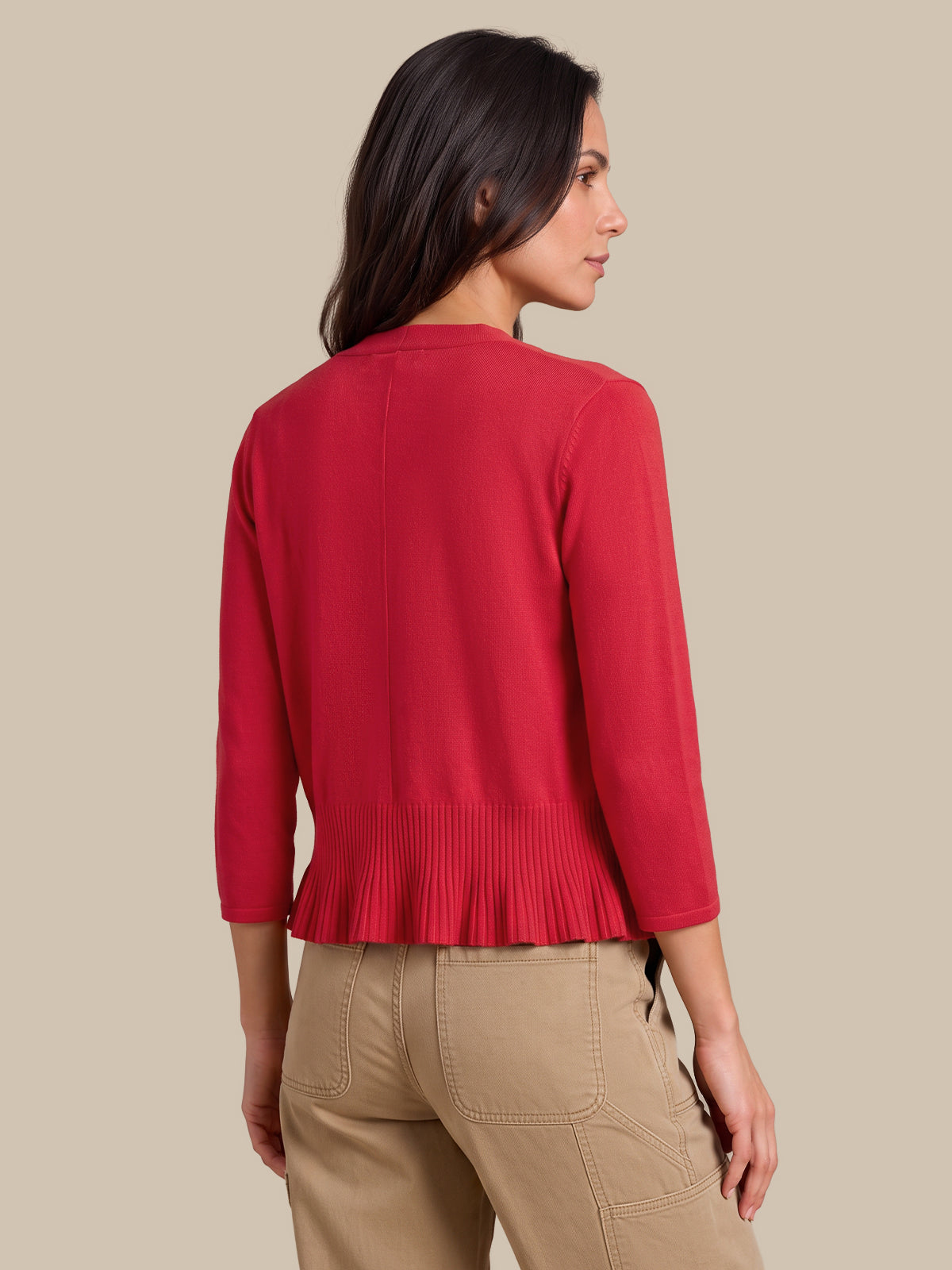 A woman shown from the back wearing a bright red knit cardigan with three-quarter sleeves and a pleated hem, paired with beige cargo pants, against a solid beige background.