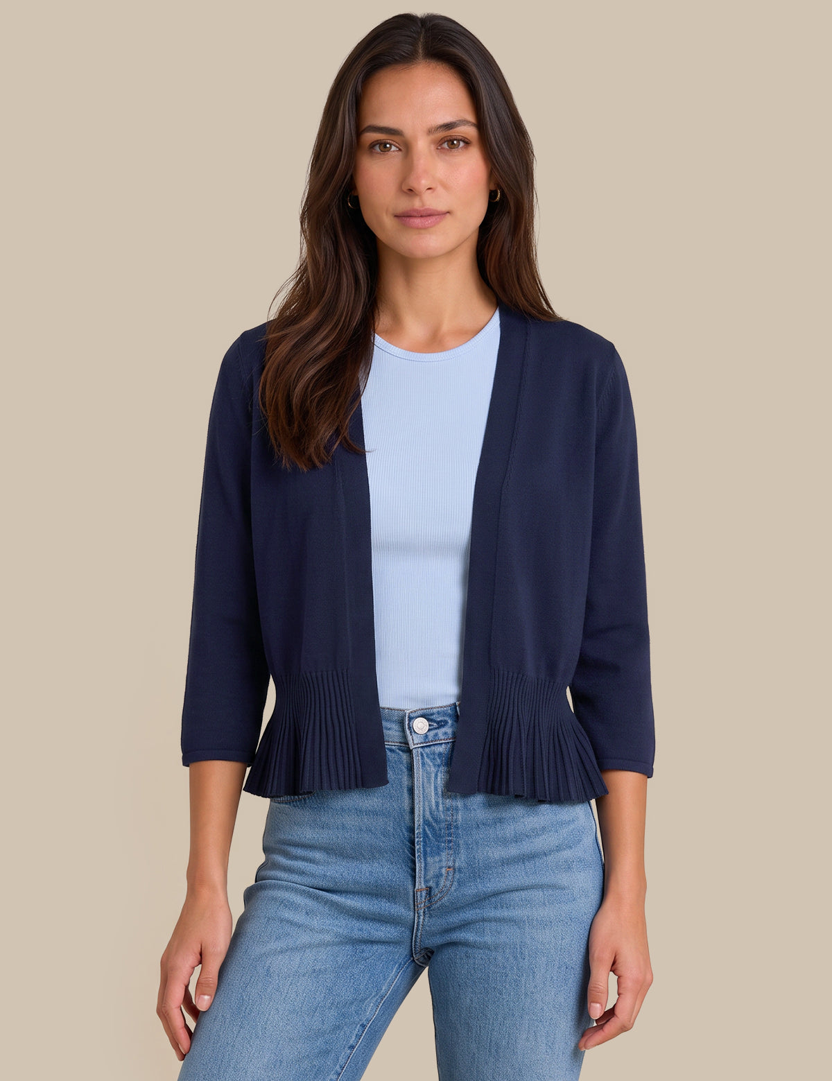 A woman modeling a navy blue open-front cardigan with pleated trim at the hem and three-quarter sleeves, worn over a light blue top and blue jeans against a solid beige background.