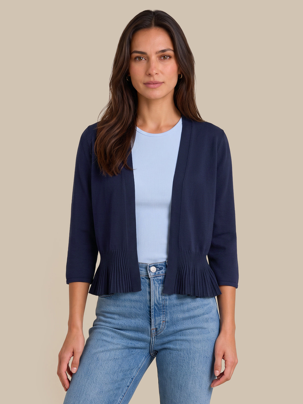 A woman modeling a navy blue open-front cardigan with pleated trim at the hem and three-quarter sleeves, worn over a light blue top and blue jeans against a solid beige background.