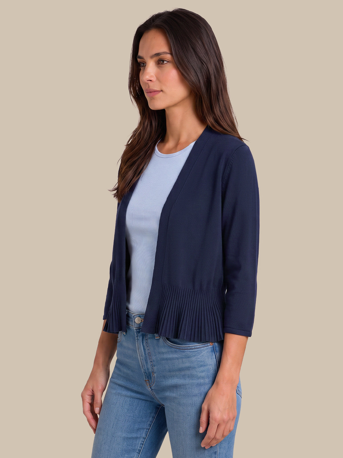 A woman wearing a navy blue open-front cardigan with pleated detailing at the hem, layered over a light blue top and paired with blue jeans, set against a solid beige background.
