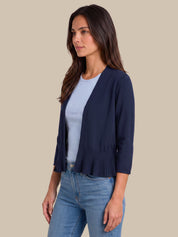 A woman wearing a navy blue open-front cardigan with pleated detailing at the hem, layered over a light blue top and paired with blue jeans, set against a solid beige background.