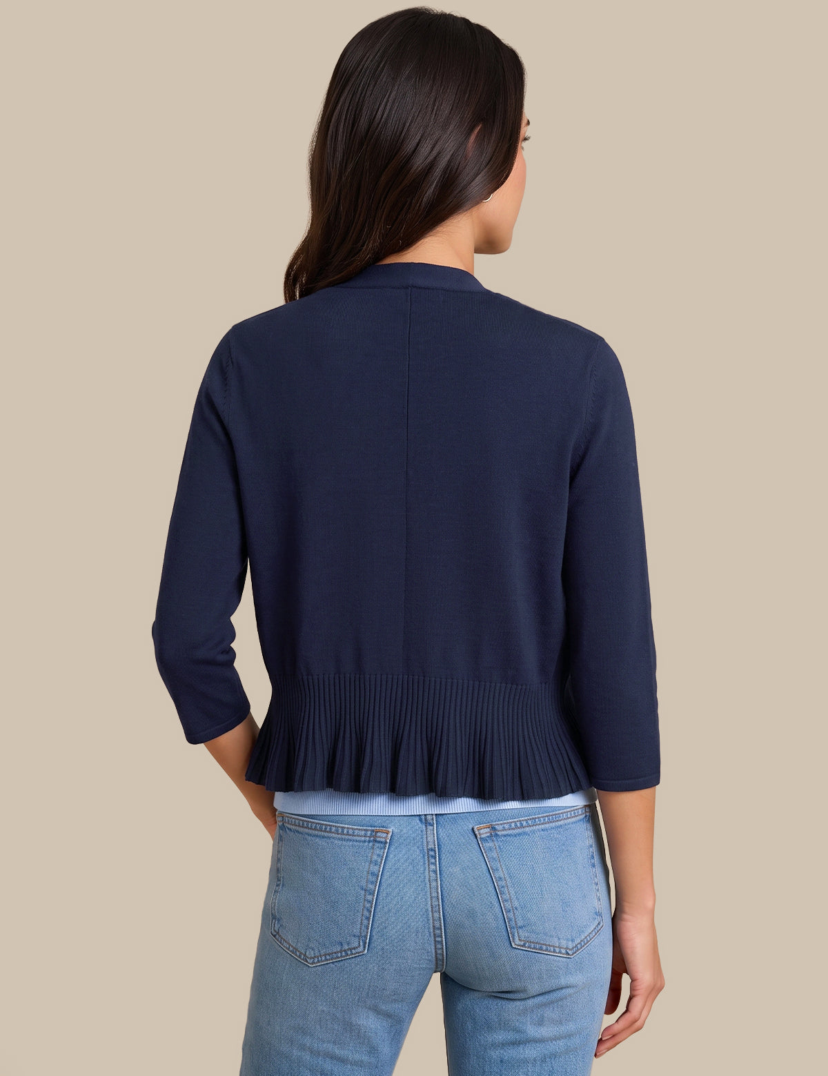 A woman shown from the back wearing a navy blue cropped cardigan with three-quarter sleeves and a pleated hem, paired with light blue jeans, against a solid beige background.