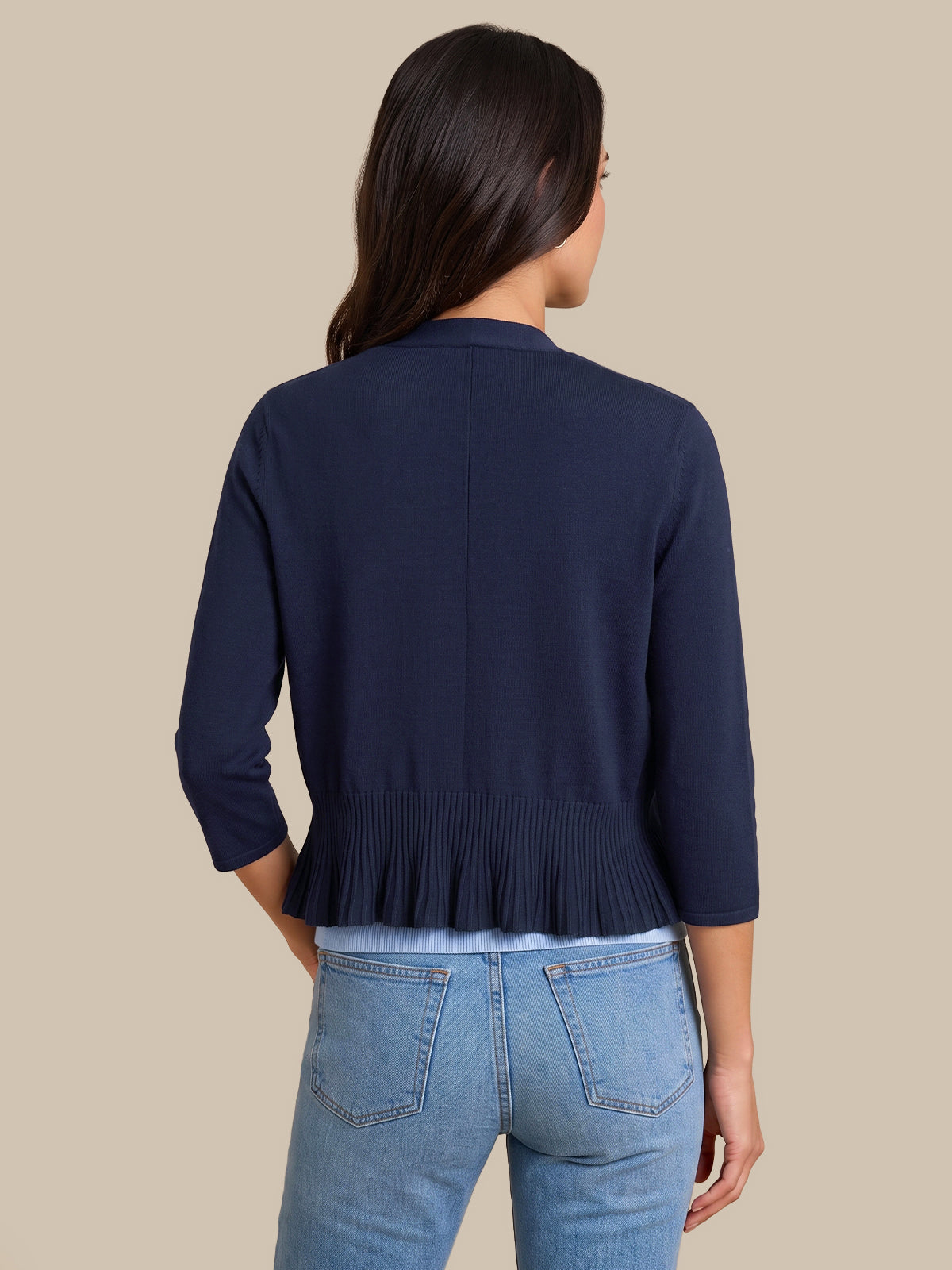 A woman shown from the back wearing a navy blue cropped cardigan with three-quarter sleeves and a pleated hem, paired with light blue jeans, against a solid beige background.