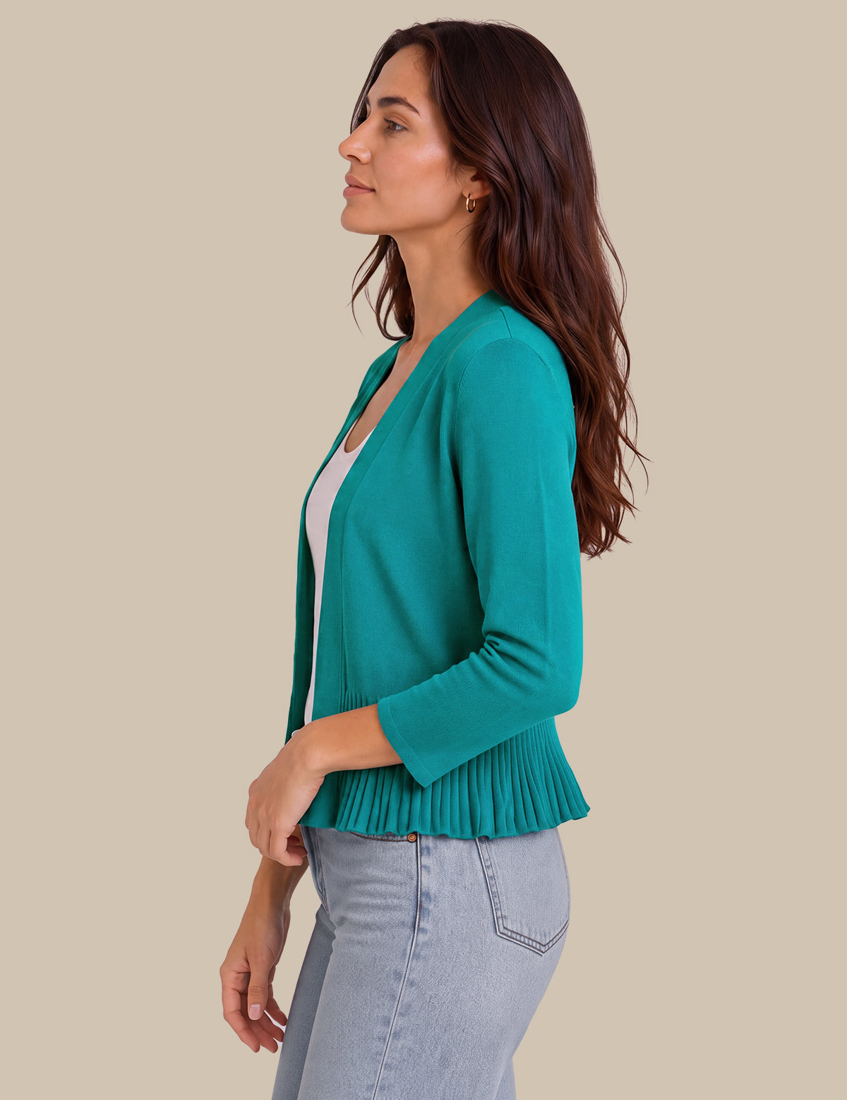 A woman in side profile wearing a teal green cardigan with three-quarter sleeves and a pleated peplum hem, styled over a white top and light wash jeans, against a solid beige background.