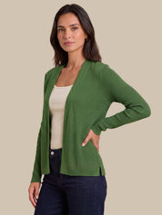 A woman modeling a green ribbed open-front cardigan sweater over a cream tank top and dark blue jeans, set against a solid beige background.