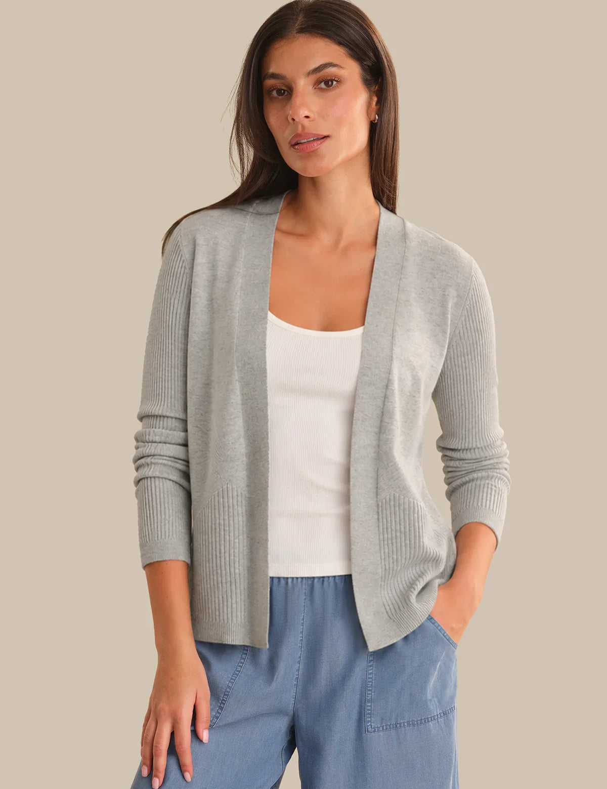 Woman wearing a gray cardigan over a white top and blue pants on a beige background