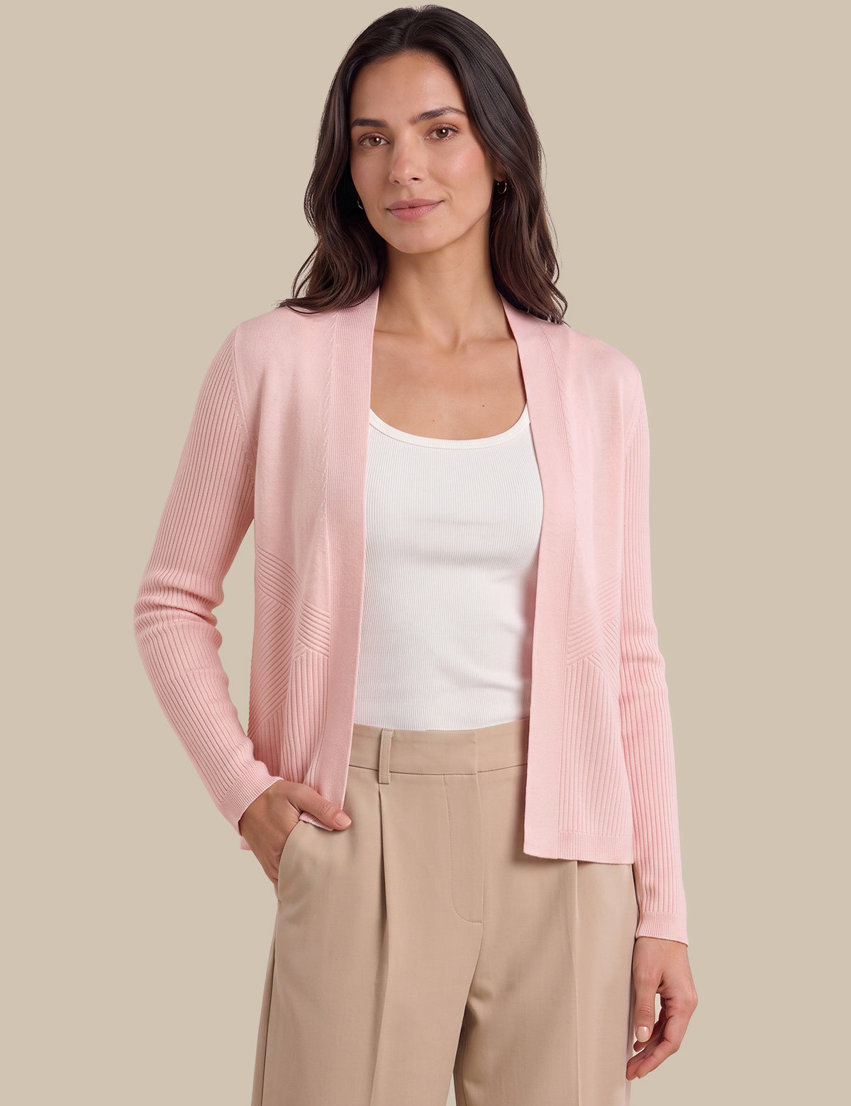 A woman modeling a light pink ribbed knit cardigan over a white tank top and beige trousers, set against a solid beige background.