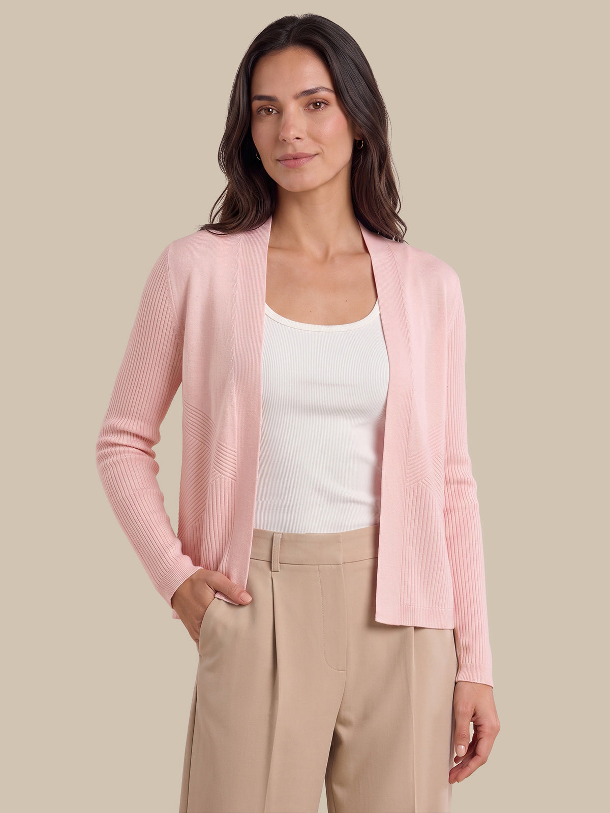 A woman modeling a light pink ribbed knit cardigan over a white tank top and beige trousers, set against a solid beige background.