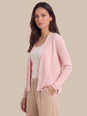 A woman modeling a light pink ribbed knit cardigan over a white tank top and beige trousers, set against a solid beige background.